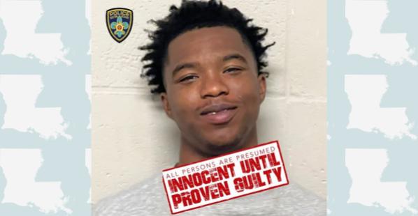 A black male from Louisiana. He has dark spikey curly hair. He is wearing a gray t-shirt and standing in front of a white brick wall.
