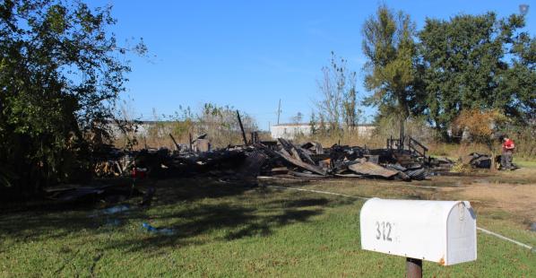 The remnants of a burned down home with a person standing next to it. There is also a mailbox in the foreground.