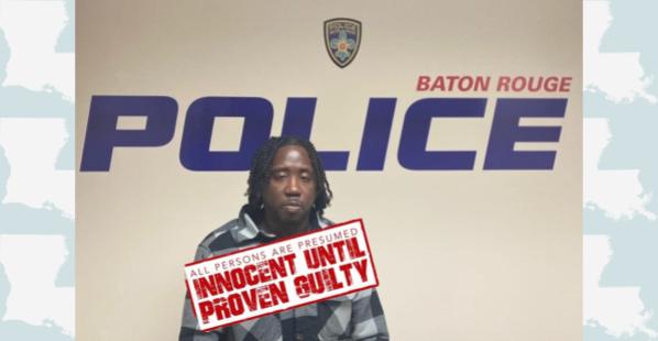 A 37-year-old black male. He has medium length, dark colored, braided hair and a thin dark mustache. He is wearing a jacket with a black, white, and gray flannel pattern. He is standing in front of a banner that reads "Baton Rouge Police."